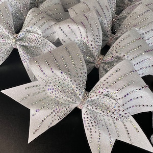 May include: Several silver glitter cheerleading bows with rhinestone accents. Each bow has a sparkling, textured finish and is adorned with rows of iridescent rhinestones. The bows are tied with a rhinestone-studded band in the center.
