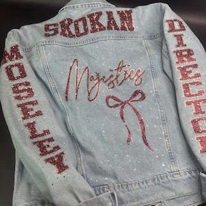 May include: A light blue denim jacket featuring red glitter lettering that reads SKOKAN on the upper back, MOSELEY on the left sleeve, and DIRECTOR on the right sleeve. The back displays the word Majestics and a red glitter bow with rhinestones.