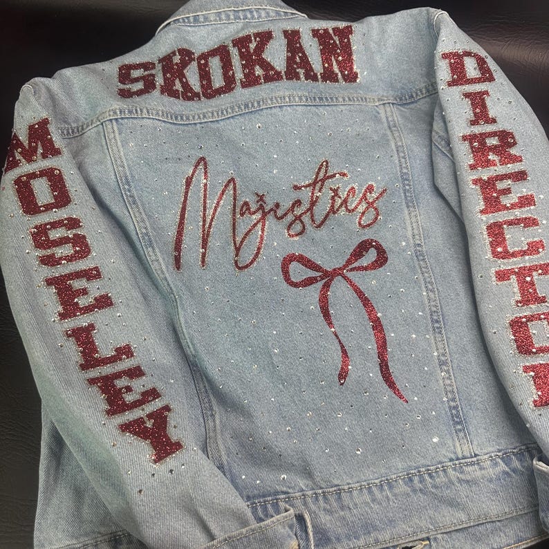 May include: A light blue denim jacket featuring red glitter lettering that reads SKOKAN on the upper back, MOSELEY on the left sleeve, and DIRECTOR on the right sleeve. The back displays the word Majestics and a red glitter bow with rhinestones.