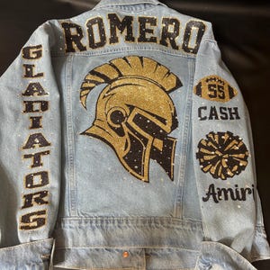 May include: A light blue denim jacket featuring a large gold and black glitter Spartan helmet on the back. The text ROMERO is at the top, GLADIATORS on the left sleeve, and 55, CASH, a pom-pom, and Amiri on the right sleeve, all accented with rhinestones.