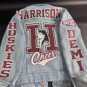 May include: A light blue denim jacket featuring red glitter lettering and accents. The back displays the name HARRISON, a large letter H with a husky dog graphic, the word Cheer, and the text HUSKIES, DEMI, and 2030 with a megaphone icon.