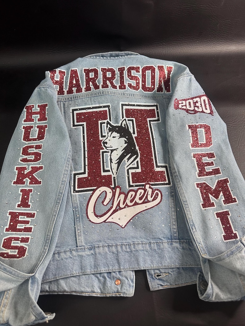 May include: A light blue denim jacket featuring red glitter lettering and accents. The back displays the name HARRISON, a large letter H with a husky dog graphic, the word Cheer, and the text HUSKIES, DEMI, and 2030 with a megaphone icon.