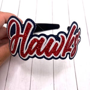 May include: A red, white, and blue glitter headband with the word "Hawks" in a cursive font.