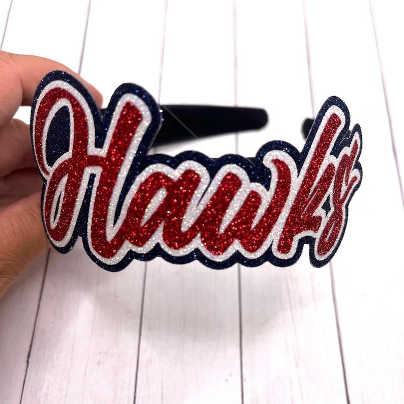 May include: A red, white, and blue glitter headband with the word "Hawks" in a cursive font.