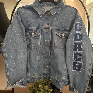 May include: A medium-wash blue denim jacket featuring a classic collar, button-front closure, and two chest pockets. The jacket is embellished with scattered white rhinestones across the front and sleeves, with the word COACH in blue letters.