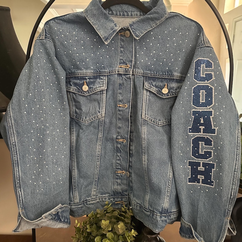 May include: A medium-wash blue denim jacket featuring a classic collar, button-front closure, and two chest pockets. The jacket is embellished with scattered white rhinestones across the front and sleeves, with the word COACH in blue letters.