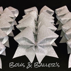 May include: A group of silver glitter cheer bows with white backing. The bows are arranged in a semi-circle with the text "Bows & Ballerz's" at the bottom of the image.