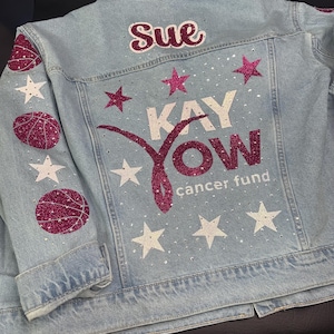 May include: A light blue denim jacket featuring the name Sue in dark pink glitter, the text KAY YOW cancer fund, and a pink glitter ribbon graphic. The back and sleeves are decorated with white and dark pink glitter stars and basketballs.