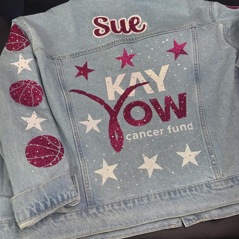 May include: A light blue denim jacket featuring the name Sue in dark pink glitter, the text KAY YOW cancer fund, and a pink glitter ribbon graphic. The back and sleeves are decorated with white and dark pink glitter stars and basketballs.