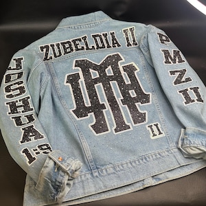 May include: A light blue denim jacket featuring black glittery lettering and a large central monogram. The back displays the text ZUBELDIA II, JOSHUA 1:9, and PMZ II. The design is accented with scattered clear rhinestones throughout the fabric.
