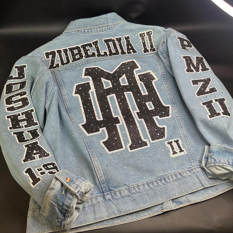 May include: A light blue denim jacket featuring black glittery lettering and a large central monogram. The back displays the text ZUBELDIA II, JOSHUA 1:9, and PMZ II. The design is accented with scattered clear rhinestones throughout the fabric.