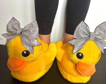 May include: A pair of bright yellow duck slippers with orange beaks and large, sparkly silver bows. The slippers are plush and have black eyes. The slippers are designed to look like cartoon ducks.
