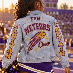 May include: A light blue denim jacket featuring purple and gold glitter lettering on the back that reads TFN, METEORS, and Cheer. The left sleeve displays the name SANIYAH in gold, and the right sleeve displays the word CAPTAIN in gold.