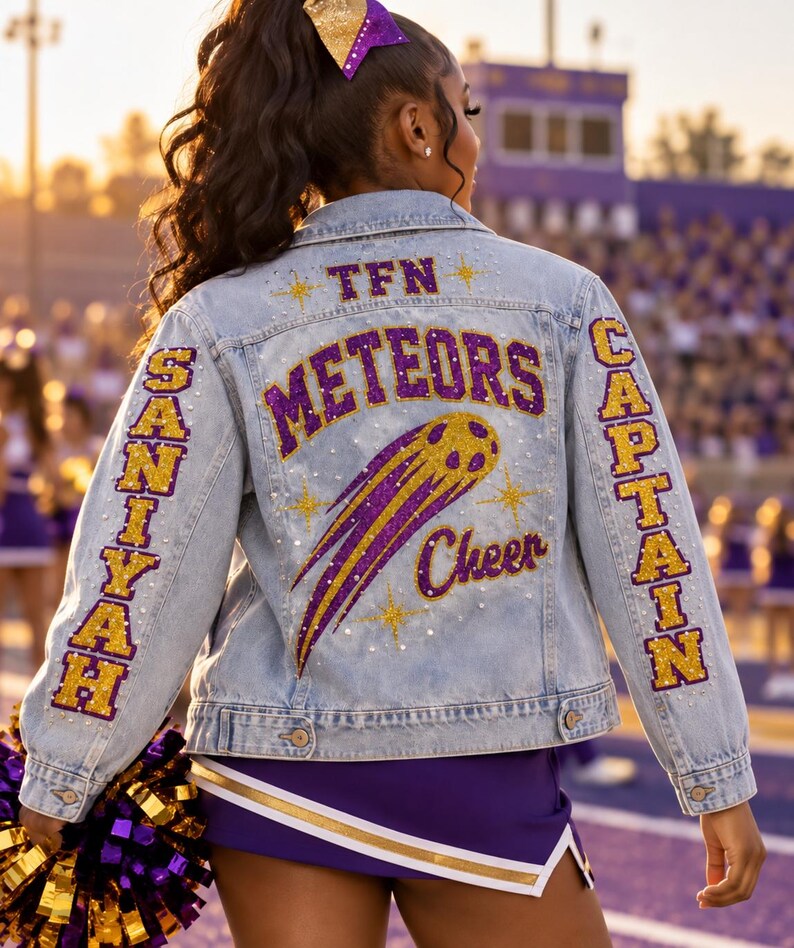 May include: A light blue denim jacket featuring purple and gold glitter lettering on the back that reads TFN, METEORS, and Cheer. The left sleeve displays the name SANIYAH in gold, and the right sleeve displays the word CAPTAIN in gold.