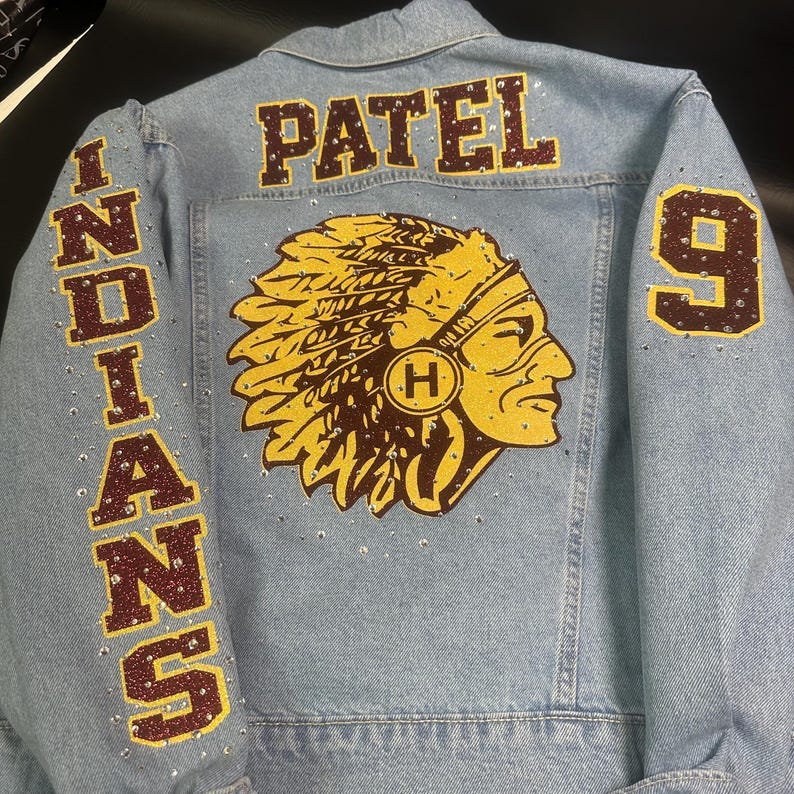 May include: A light blue denim jacket featuring the name PATEL in dark red letters across the top back. The centre back displays a yellow and dark red graphic of a person wearing a headdress. The left sleeve says INDIANS and the right sleeve has the number 9, all accented with clear rhinestones.