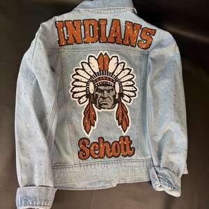 May include: A light blue denim jacket featuring the word INDIANS in orange glitter lettering at the top, a central graphic of a person in a feathered headdress, and the word Schott in orange glitter lettering at the bottom, with rhinestones.