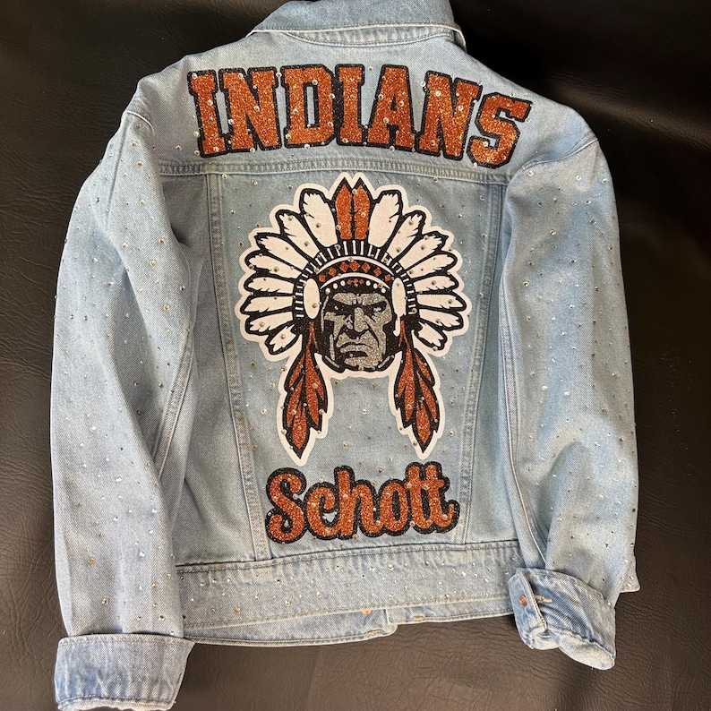 May include: A light blue denim jacket featuring the word INDIANS in orange glitter lettering at the top, a central graphic of a person in a feathered headdress, and the word Schott in orange glitter lettering at the bottom, with rhinestones.