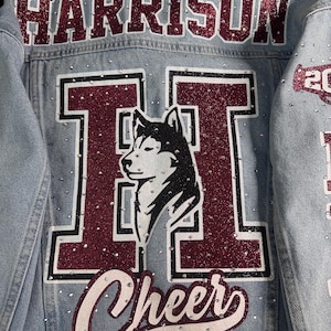 May include: A light blue denim jacket featuring the name HARRISON in maroon glitter letters across the top. A large maroon letter H with a white husky dog silhouette is centered on the back, with the word Cheer below it, all accented by rhinestones.
