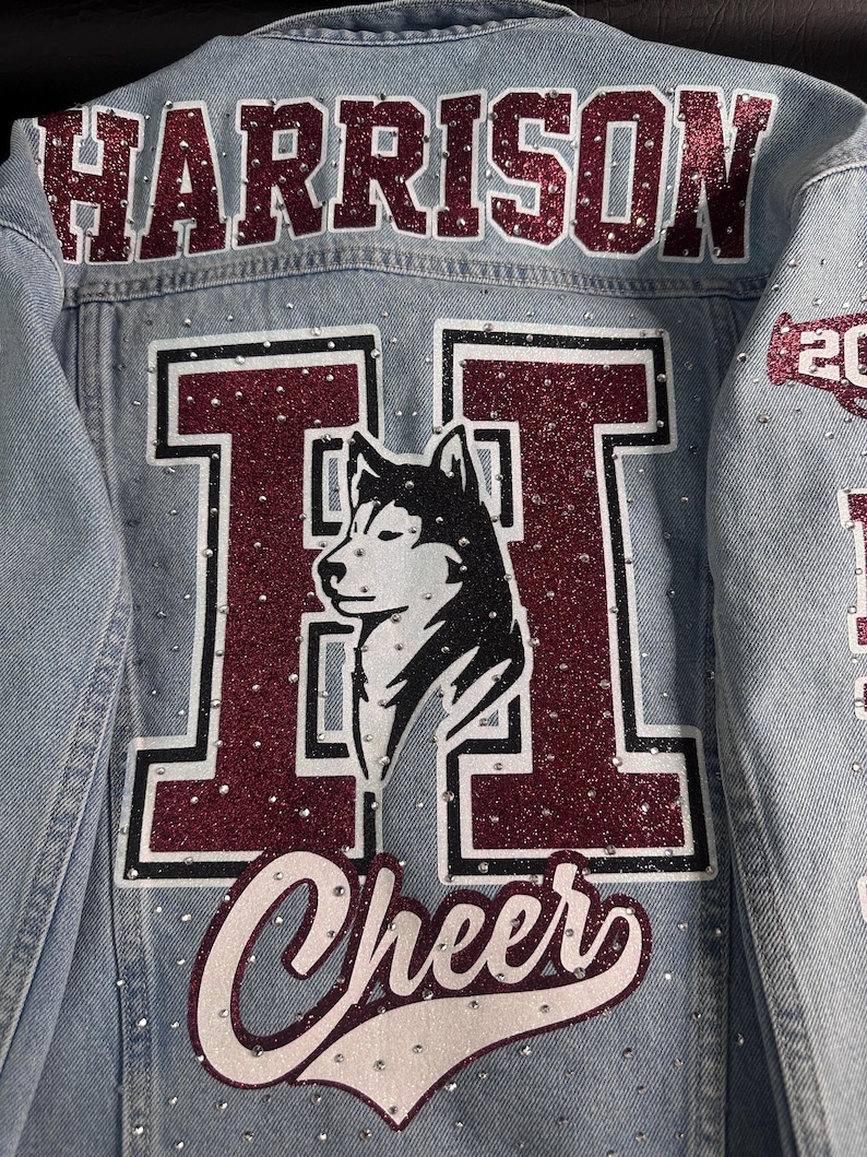 May include: A light blue denim jacket featuring the name HARRISON in maroon glitter letters across the top. A large maroon letter H with a white husky dog silhouette is centered on the back, with the word Cheer below it, all accented by rhinestones.