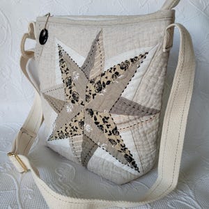 May include: Cream-colored quilted crossbody bag with a large star design. The star is composed of various fabric patches in shades of cream, tan, and black floral patterns. The bag has a long, adjustable strap and a black button closure.
