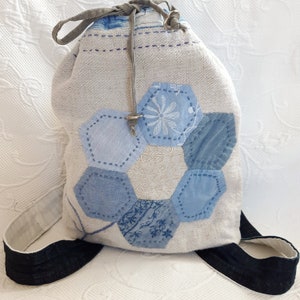 Könnte beinhalten: Eine weiße Kordeltasche mit einem blauen und weißen Patchwork-Muster. Die Tasche hat einen langen Riemen und ist aus einem weichen, strukturierten Stoff gefertigt.