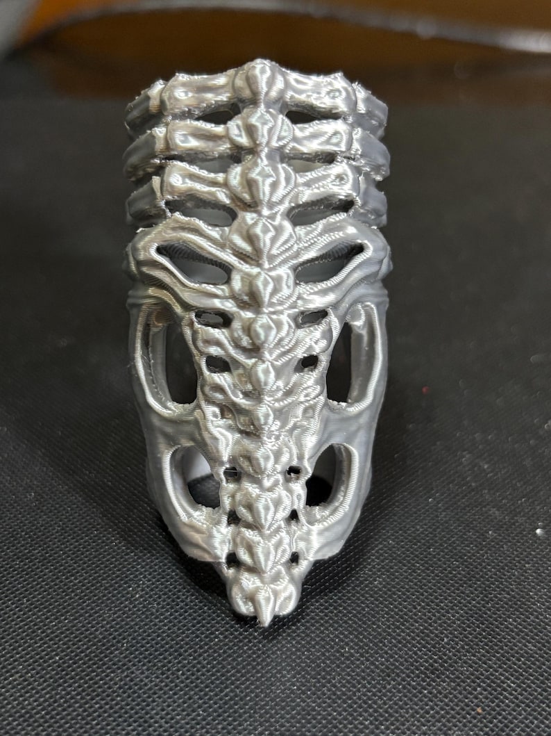 May include: A silver-colored ring with a detailed skeletal rib cage design. The ring features a three-dimensional, textured appearance, with open spaces mimicking the spaces between ribs. The ring is likely made of metal and is designed to be worn on a finger.
