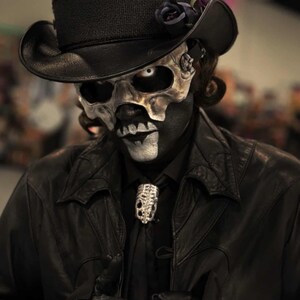 May include: A person in a black leather jacket, black shirt, and a skull mask. They wear a black top hat with a dark rose and feather. A silver skull-shaped brooch adorns the black tie. The makeup creates a striking contrast.
