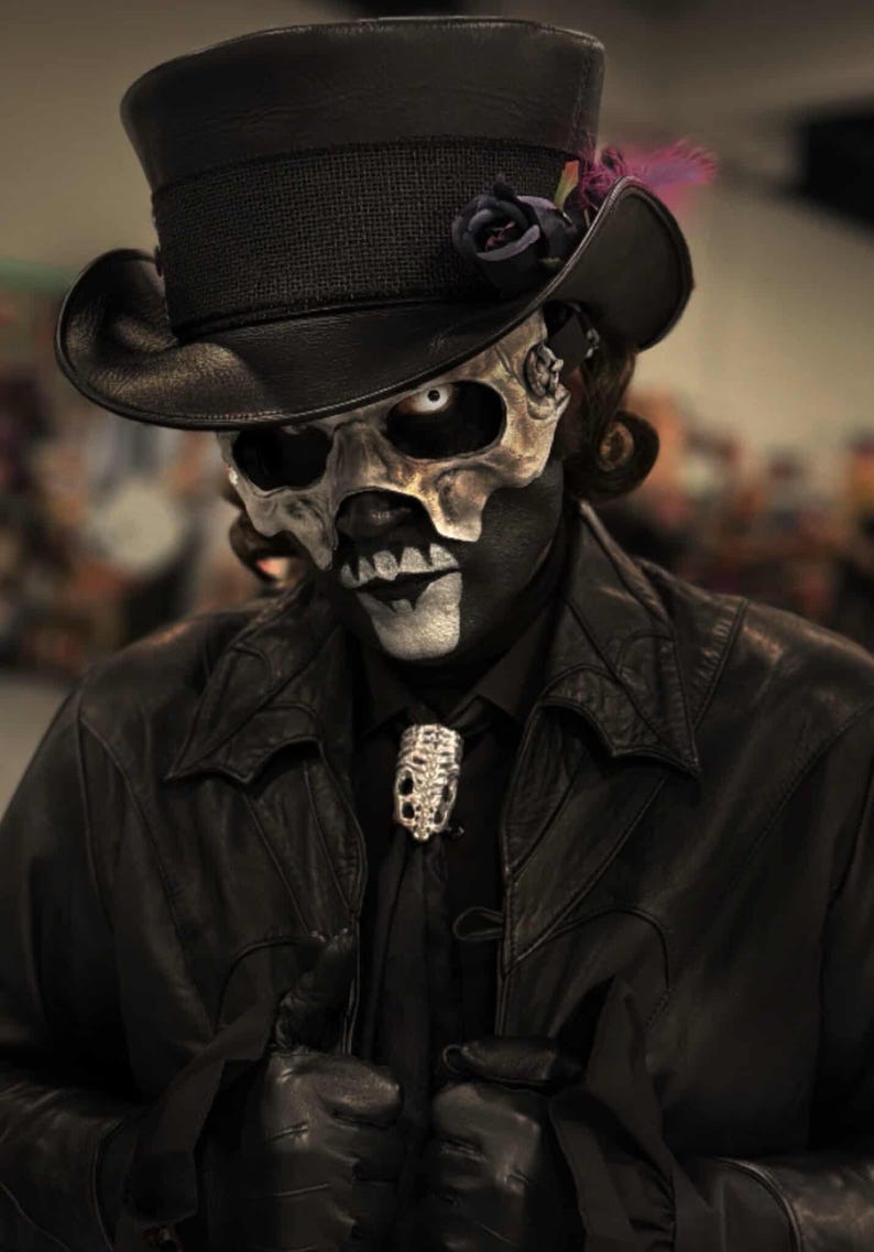 May include: A person in a black leather jacket, black shirt, and a skull mask. They wear a black top hat with a dark rose and feather. A silver skull-shaped brooch adorns the black tie. The makeup creates a striking contrast.