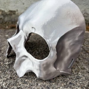 May include: A silver-colored skull mask with eye and nose openings. The mask has a textured surface and is made of a solid material. The mask is designed to be worn on the face and is likely for costume or decorative purposes.