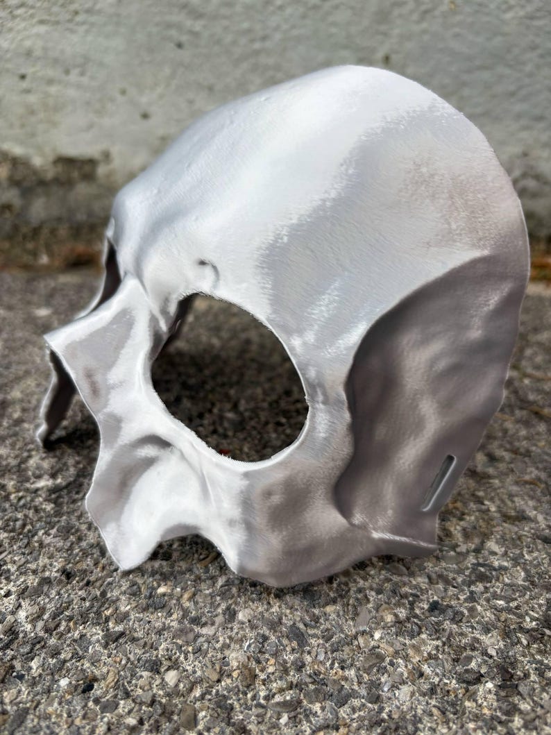 May include: A silver-colored skull mask with eye and nose openings. The mask has a textured surface and is made of a solid material. The mask is designed to be worn on the face and is likely for costume or decorative purposes.