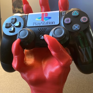 May include: A red 3D printed hand holding a black PlayStation 4 controller. The controller has the PlayStation logo and buttons.