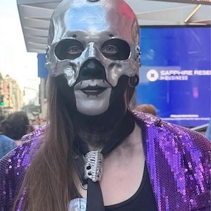 May include: A person wearing a silver skull mask with black accents, a black tie with a skull-shaped tie clip, and a purple sequined jacket. The background includes signs for Penn Station and Madison Square Garden.