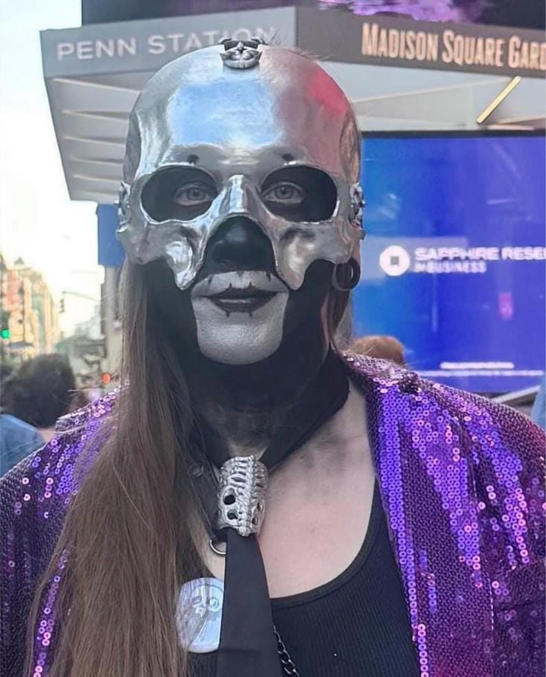 May include: A person wearing a silver skull mask with black accents, a black tie with a skull-shaped tie clip, and a purple sequined jacket. The background includes signs for Penn Station and Madison Square Garden.