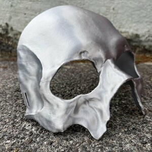 May include: A silver-colored skull mask with a large eye opening and detailed facial features. The mask is made of a metallic material and has a textured surface. The mask is designed to be worn and is likely for costume or decorative purposes.