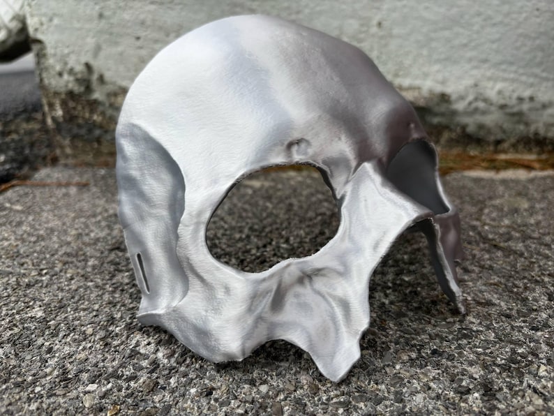 May include: A silver-colored skull mask with a large eye opening and detailed facial features. The mask is made of a metallic material and has a textured surface. The mask is designed to be worn and is likely for costume or decorative purposes.