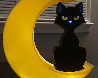 以下が含まれることがあります： 黄色い三日月の上に座っている黒い猫のシルエット。猫は青い耳と黄色い目をしています。月は内側から光っていて、温かい光を放っています。