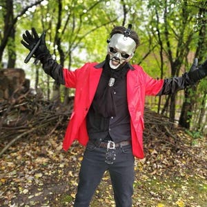 May include: A person in a red jacket, black shirt, and black pants, wearing a silver skull mask and black gloves, standing in a forest setting. The person is wearing a black belt with a silver buckle.