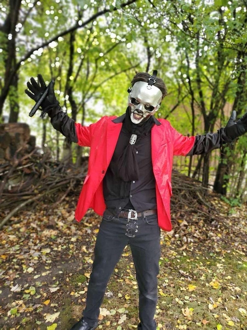 May include: A person in a red jacket, black shirt, and black pants, wearing a silver skull mask and black gloves, standing in a forest setting. The person is wearing a black belt with a silver buckle.