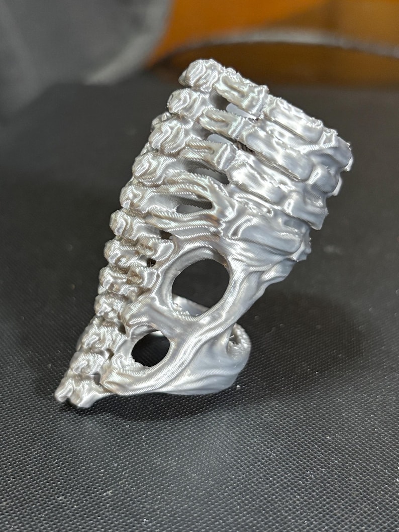 May include: A silver-colored, 3D-printed ring with a unique, organic design. The ring features a textured surface and an open, skeletal structure, with two large circular openings. The overall shape is asymmetrical and artistic.