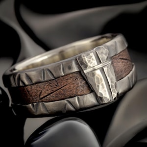 May include: A wide band ring featuring a dark brown wood inlay centered between two brushed silver edges. The silver edges have a textured, hammered finish with triangular notches, and a prominent hammered silver cross emblem is set on the front.