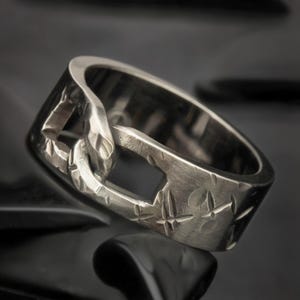 May include: A silver-toned metal ring featuring a wide band with a textured, brushed finish. The design incorporates a central cutout section that resembles an interlocking chain link, accented by small, engraved star-shaped patterns on the surface.