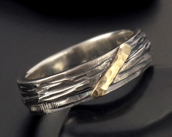 Puede incluir: Un anillo de banda de plata con un acabado oscuro oxidado y una superficie texturizada y rugosa. Una incrustación diagonal única de color oro martillado cruza la banda, proporcionando un contraste suave y reflectante con la textura oscura.
