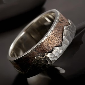 May include: A wide band ring featuring a two-tone design with a polished silver interior and a textured exterior. The outer surface combines a dark, weathered brown finish with a bright, hammered silver section that has a jagged, mountain-like edge.
