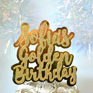 May include: A gold glitter cake topper with the words "Sofia's Golden Birthday" in a cursive font. The topper is placed on top of a white frosted cake. The background has a blurred, sparkly effect.