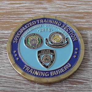 Op de afbeelding: Een ronde goudkleurige challenge coin met een lichtblauw midden en een donkerblauwe buitenring. De tekst luidt SPECIALIZED TRAINING SECTION en TRAINING BUREAU. Het bevat drie verschillende politie-emblemen en het jaartal 2015.