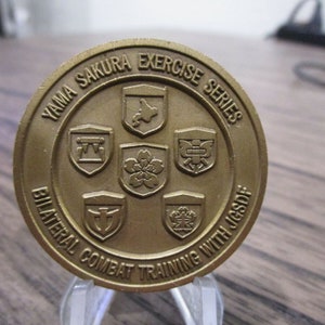 以下が含まれることがあります： 縁に「Yama Sakura Exercise Series」と「Bilateral Combat Training with JGSDF」の文字が刻まれた金色のコイン。コインの中央には、異なるシンボルが描かれた5つの盾がある。
