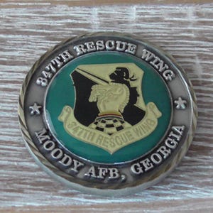 Puede incluir: Una moneda redonda de color plateado con el emblema del 347th Rescue Wing. El emblema es un escudo con fondo verde, una silueta negra y el texto "347th Rescue Wing, Moody AFB, Georgia."