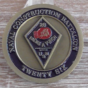Pode incluir: Medalha redonda do Naval Construction Battalion, de cor bronze. A medalha apresenta o texto "Naval Construction Battalion Twenty Six" ao redor da borda. O centro tem um losango com uma luva de boxe vermelha e as palavras "Packs a Punch".