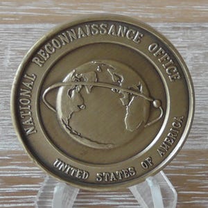 Może przedstawiać: Brązowa moneta z napisem "National Reconnaissance Office, United States of America" i globusem z orbitą wokół niego.