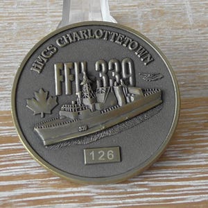Może przedstawiać: Okrągły, brązowy medal z napisem "HMCS CHARLOTTETOWN" i wizerunkiem okrętu wojennego. Medal zawiera również liść klonu oraz numery "FFH 339" i "126".